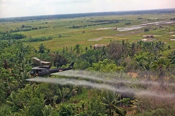 Photographie de la guerre du Viêt Nam représentant un hélicoptère américain en plein épandage d'agent orange (le célèbre défoliant produit par Monsanto) sur les campagnes vietnamiennes - considérablement et durablement contaminées par ce dernier.